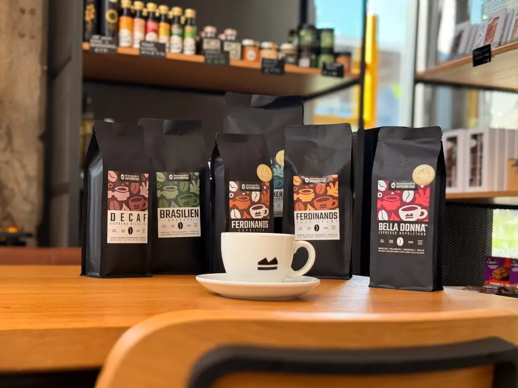 5 Tüten Kaffee stehend auf dem Tisch, davor eine Tasse mit Genusshöfe Logo