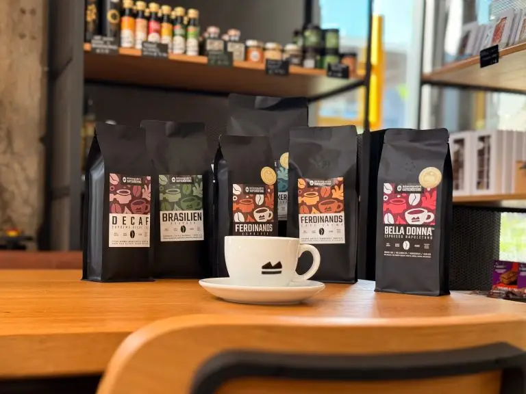 5 Tüten Kaffee stehend auf dem Tisch, davor eine Tasse mit Genusshöfe Logo