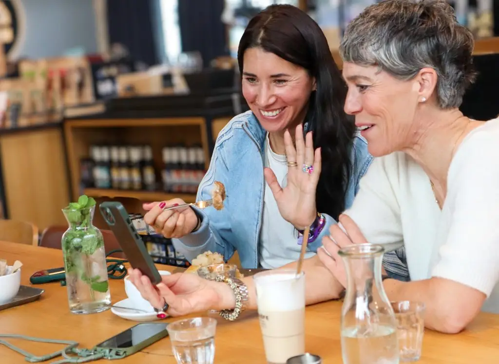 2 Frauen sitzen an einem Tisch in einem Kaffeehaus und haben einen Videocall am Handy. Eine Frau isst eine Zimtschnecke.